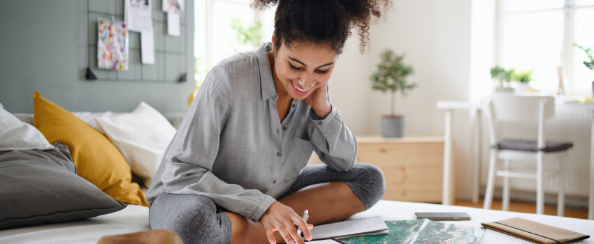 Une femme souriante assise sur son lit en train de regarder sur son cahier de notes